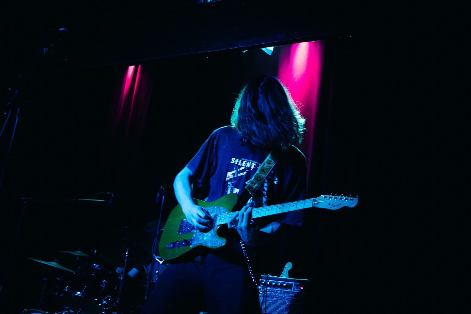 A guitarist performs on stage illuminated by vibrant pink and blue lighting during a concert performance.