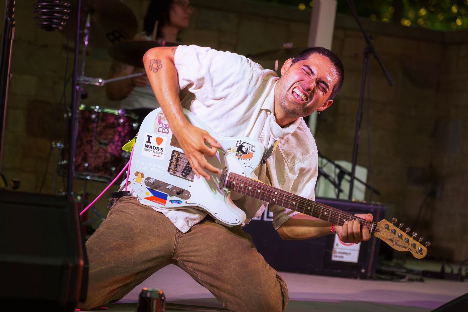 Energetic guitarist performing on stage, wearing a white t-shirt with stickers, playing electric guitar at outdoor concert.
