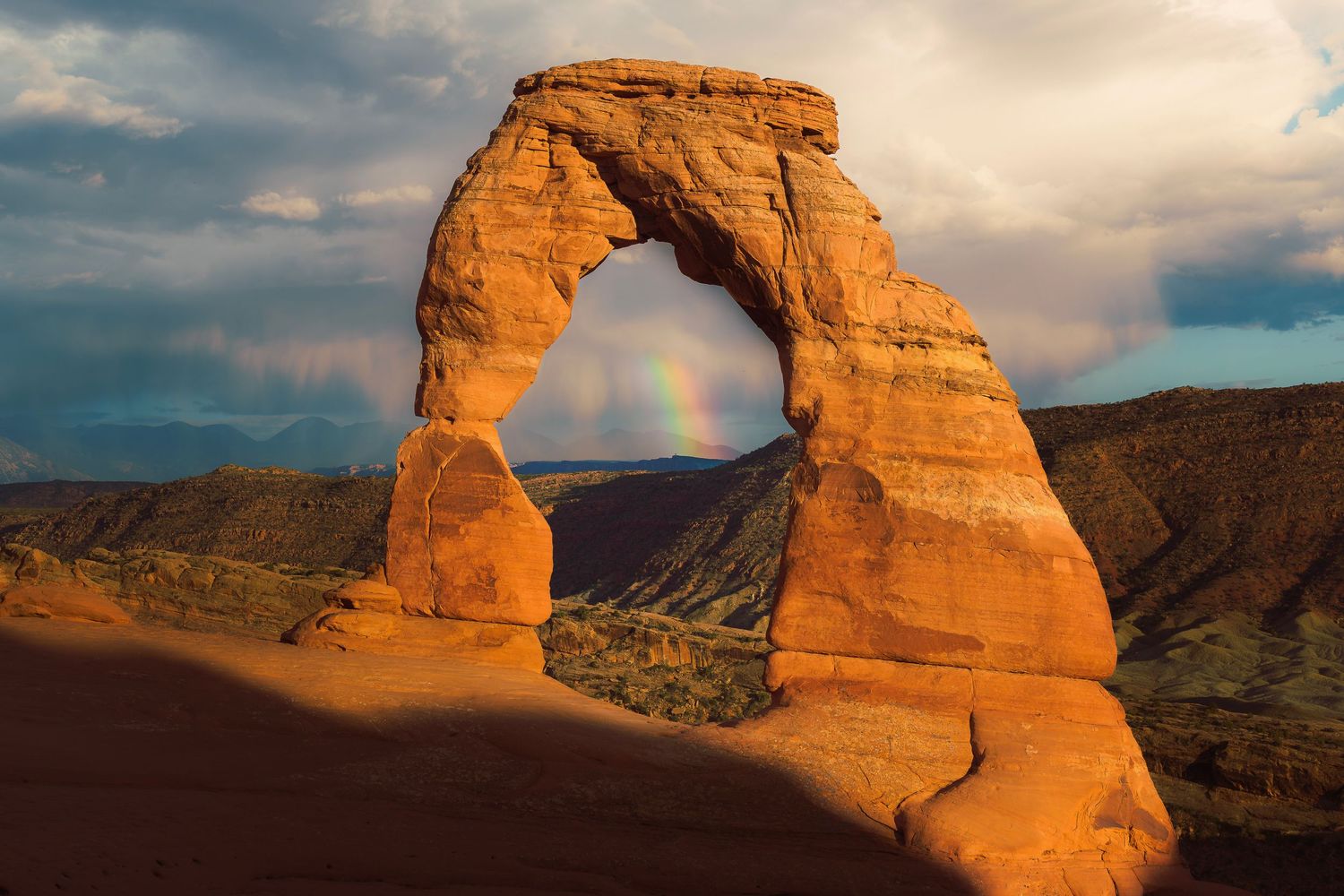 Delicate Arch at sunset with rainbow visible through sandstone opening in Arches National Park, Utah.