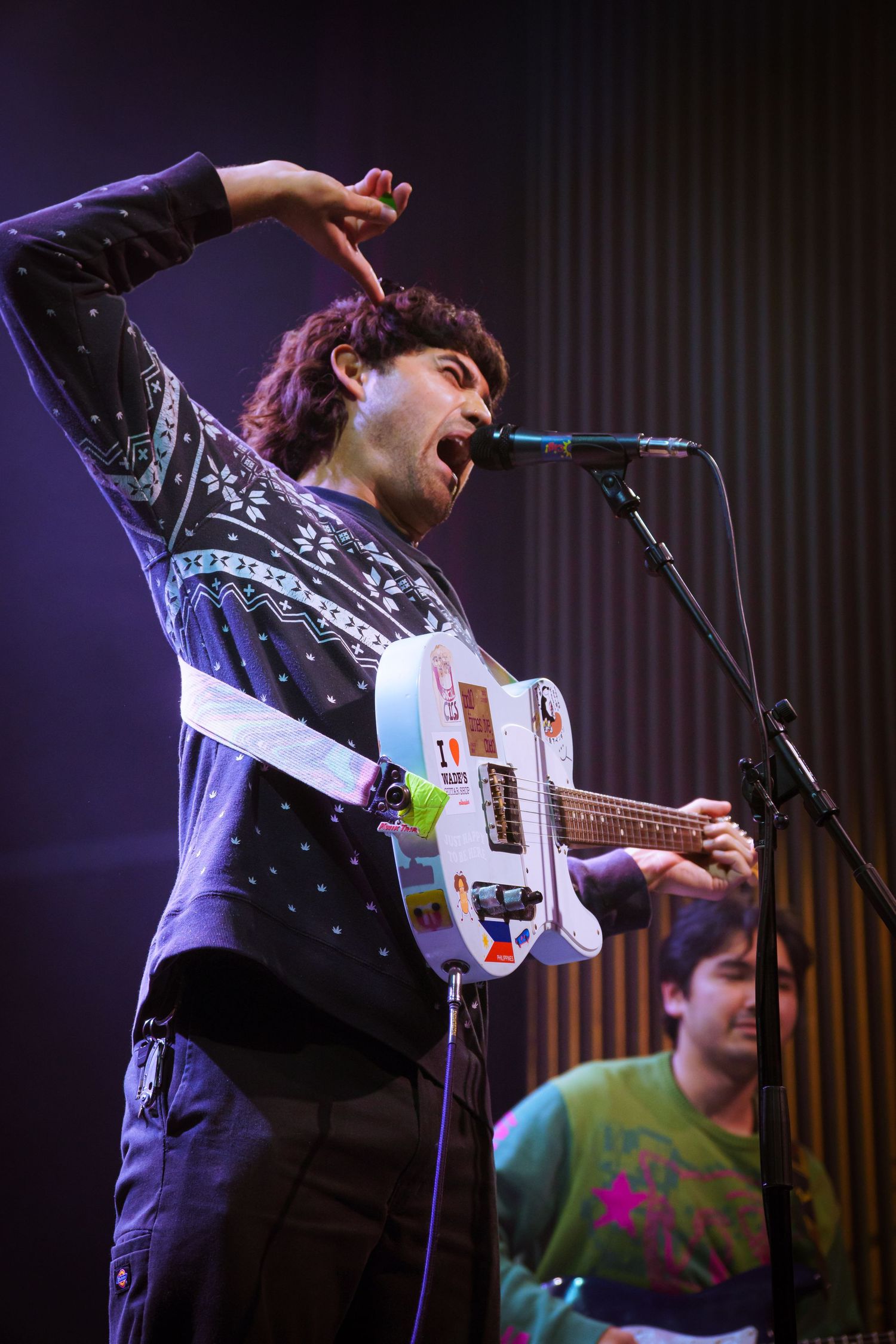 Musician performs energetically on stage with an electric guitar under purple stage lighting.