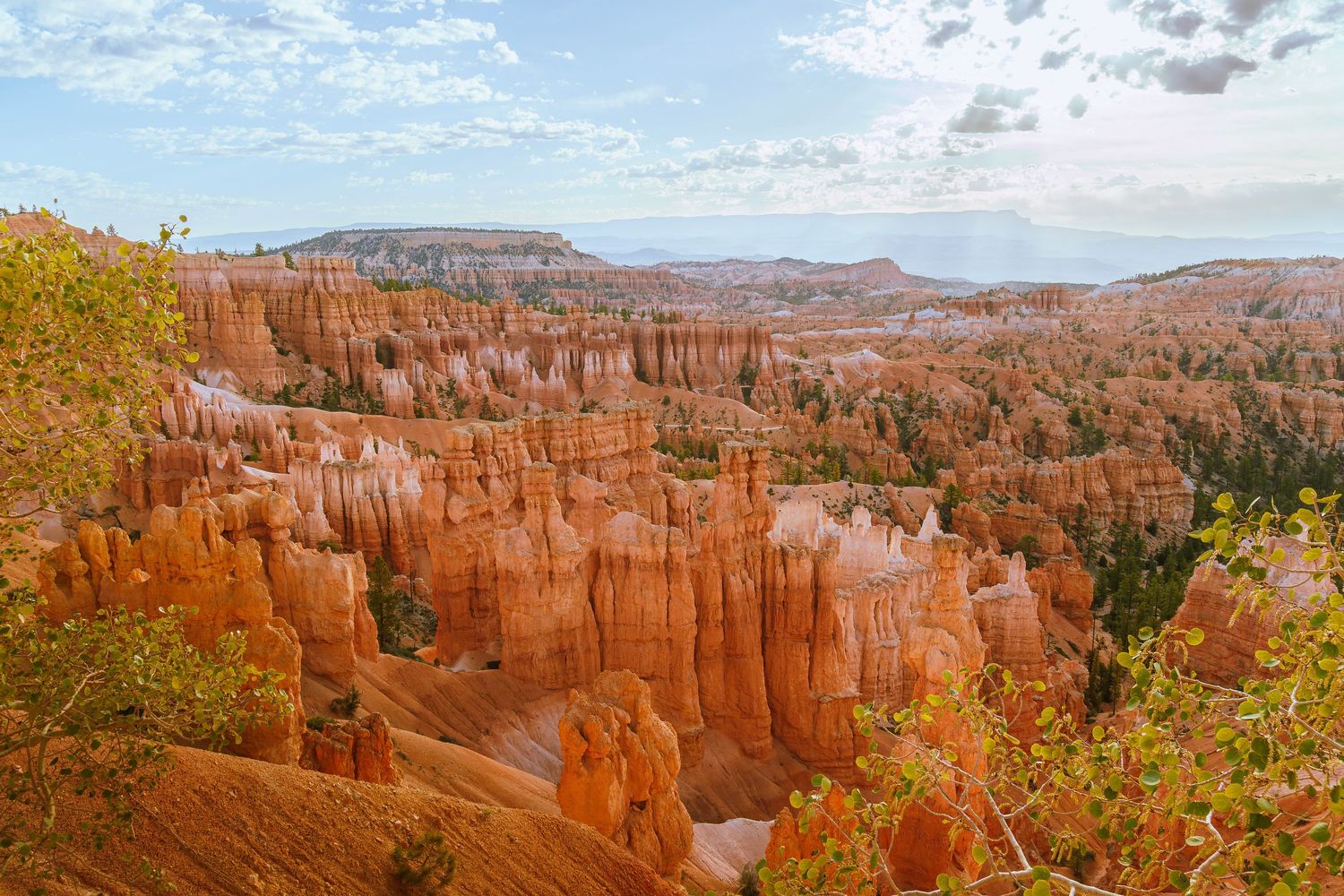 Panoramic view of Bryce Canyon hoodoos, orange rock spires rising from red Utah desert landscape under partly cloudy sky.