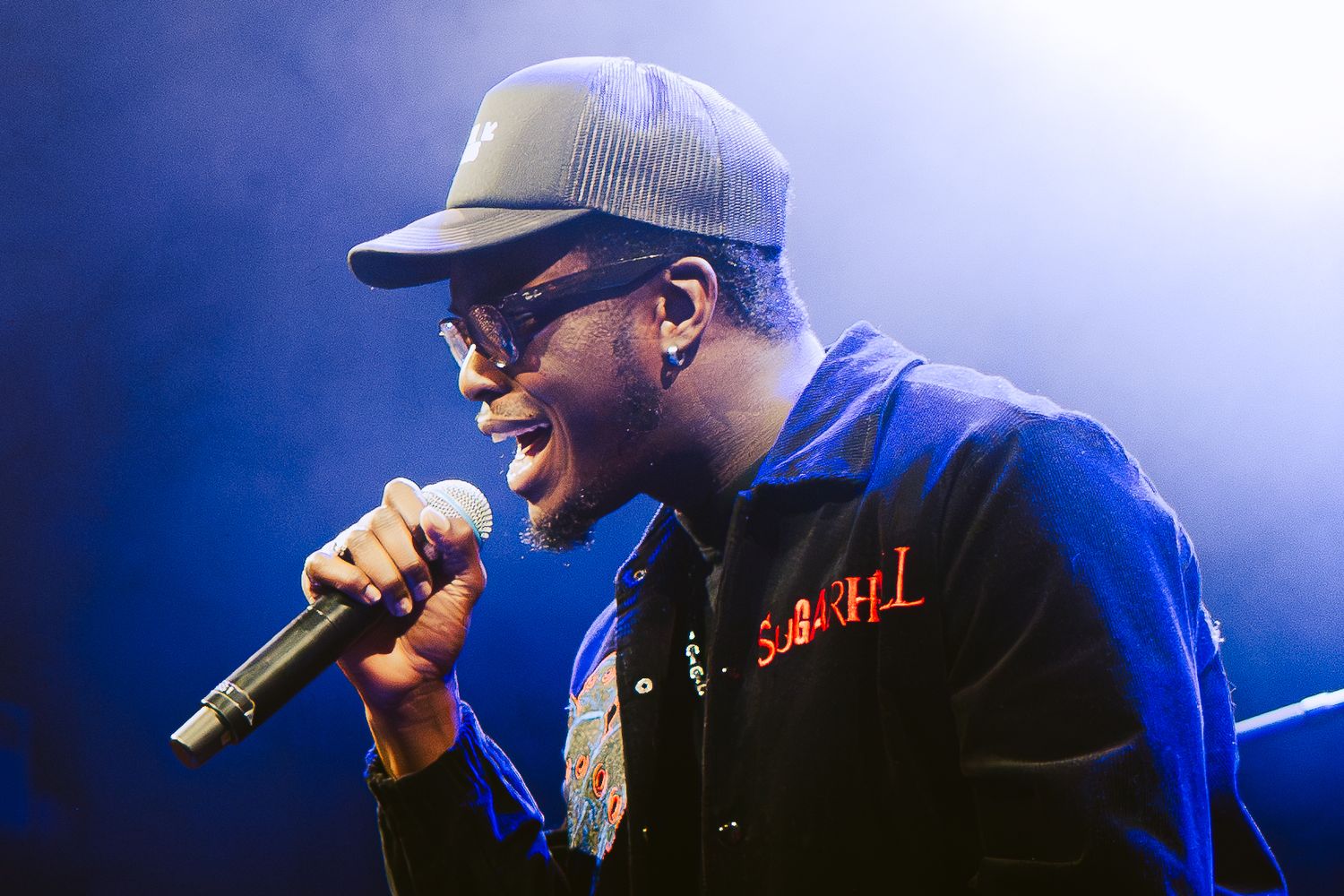 A performer in a blue shirt and baseball cap sings passionately into a microphone on stage with blue lighting.