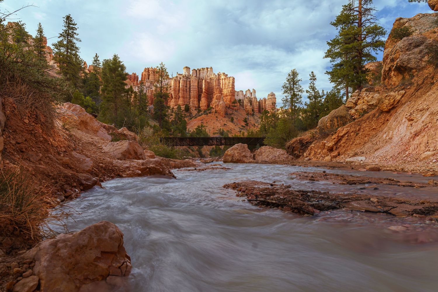 Red rock canyon with rushing river, bridge, and pine trees under dramatic cloudy sky in Bryce Canyon, Utah.