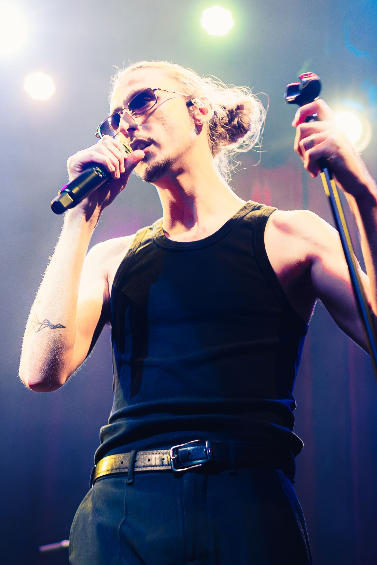 Performer in black tank top and sunglasses singing on stage with microphones under bright concert lighting.
