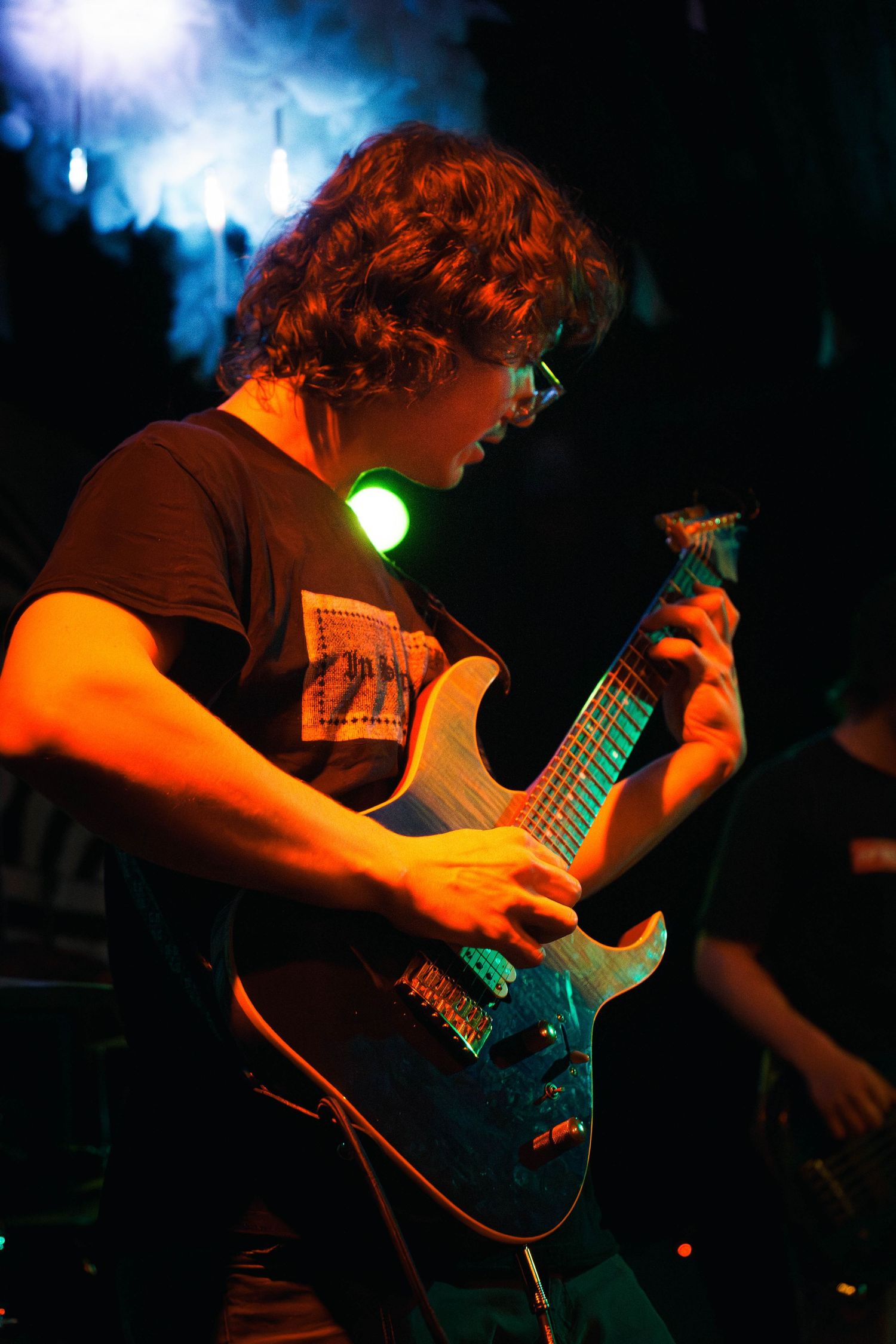 Guitarist performs on stage in dramatic orange lighting while playing an electric guitar during a live concert performance.