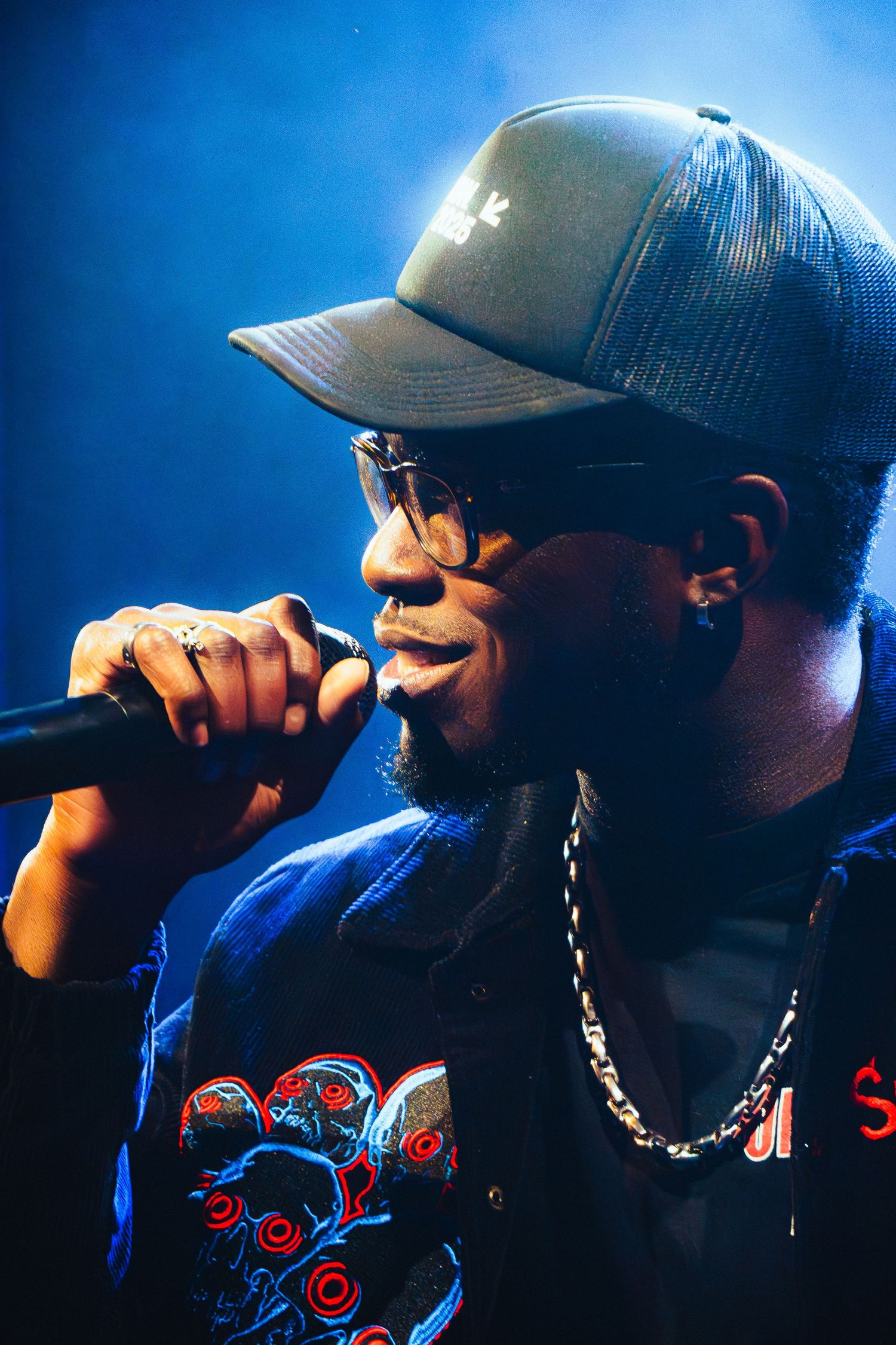 Performer in baseball cap and sunglasses singing into microphone against blue background during concert performance.