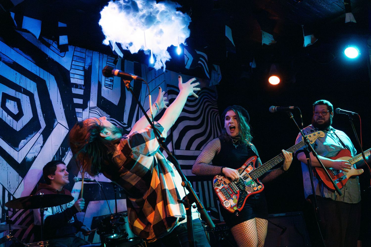 Rock band performs energetically on stage with dramatic lighting and geometric patterns on backdrop.