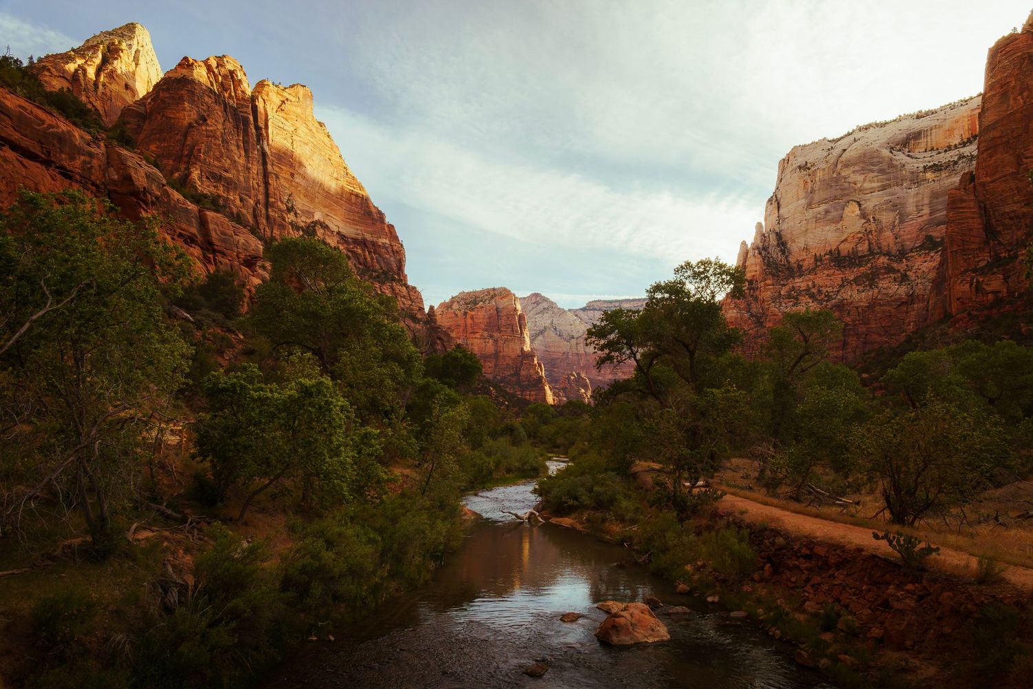Red rock canyon with a winding river, lush green trees, and dramatic cliffs under a partly cloudy sky in Zion National Park.
