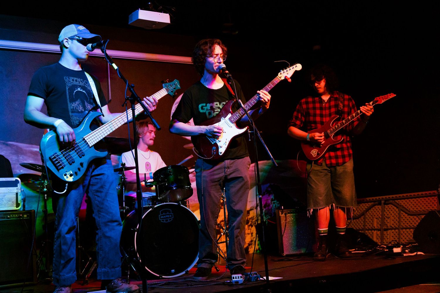 Band performing live on stage with multiple guitarists and a drummer in the background.