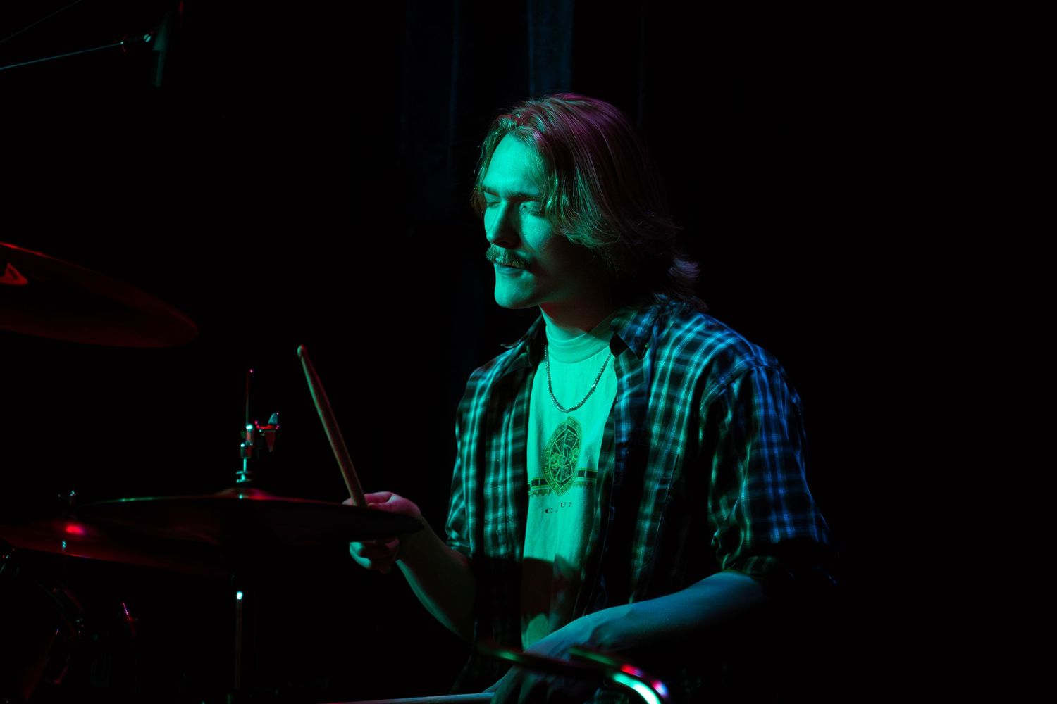 Performer in plaid shirt plays guitar on dimly lit stage with dramatic green and red lighting effects.