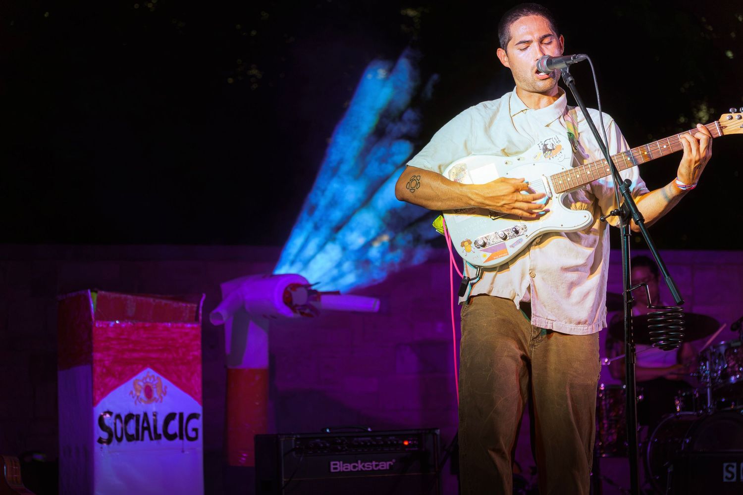 Guitarist performing on stage at night with a Stratocaster guitar near a Social Cig sign under colorful lights.