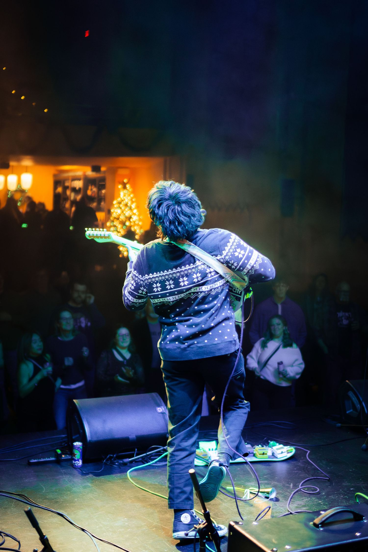 A performer wearing a blue patterned sweater plays guitar on stage at a dimly lit concert venue with a crowd visible.