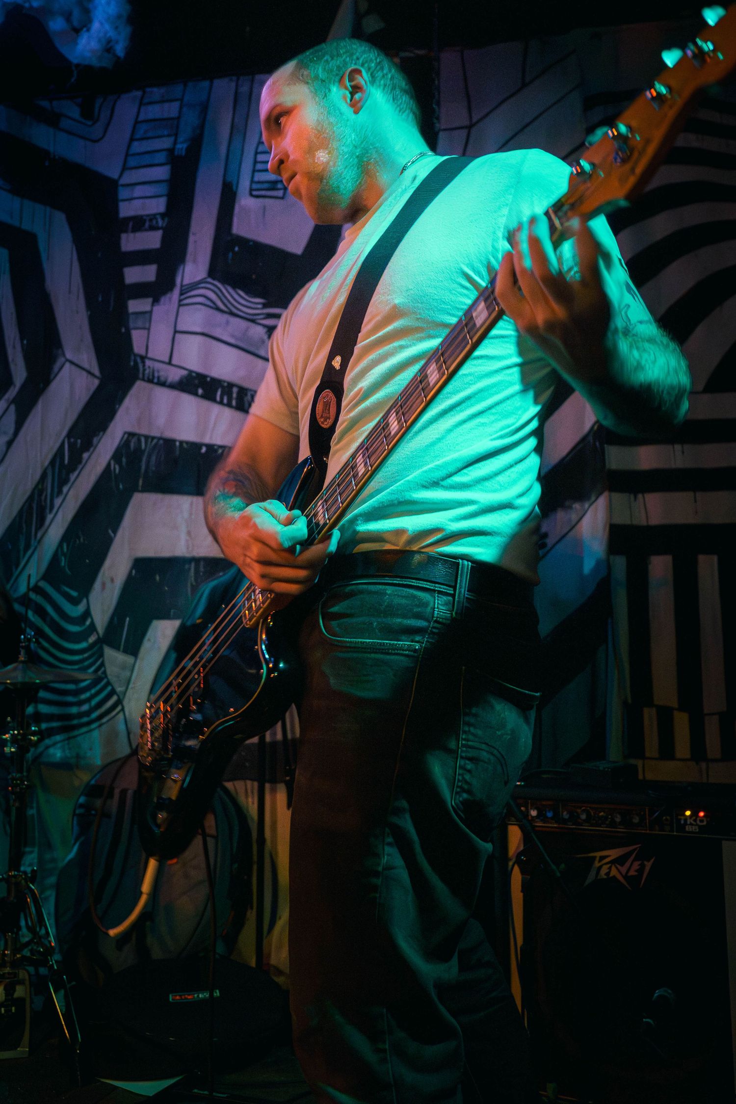 Bass guitarist performing on stage under vibrant turquoise lighting with dramatic shadows and geometric backdrop.