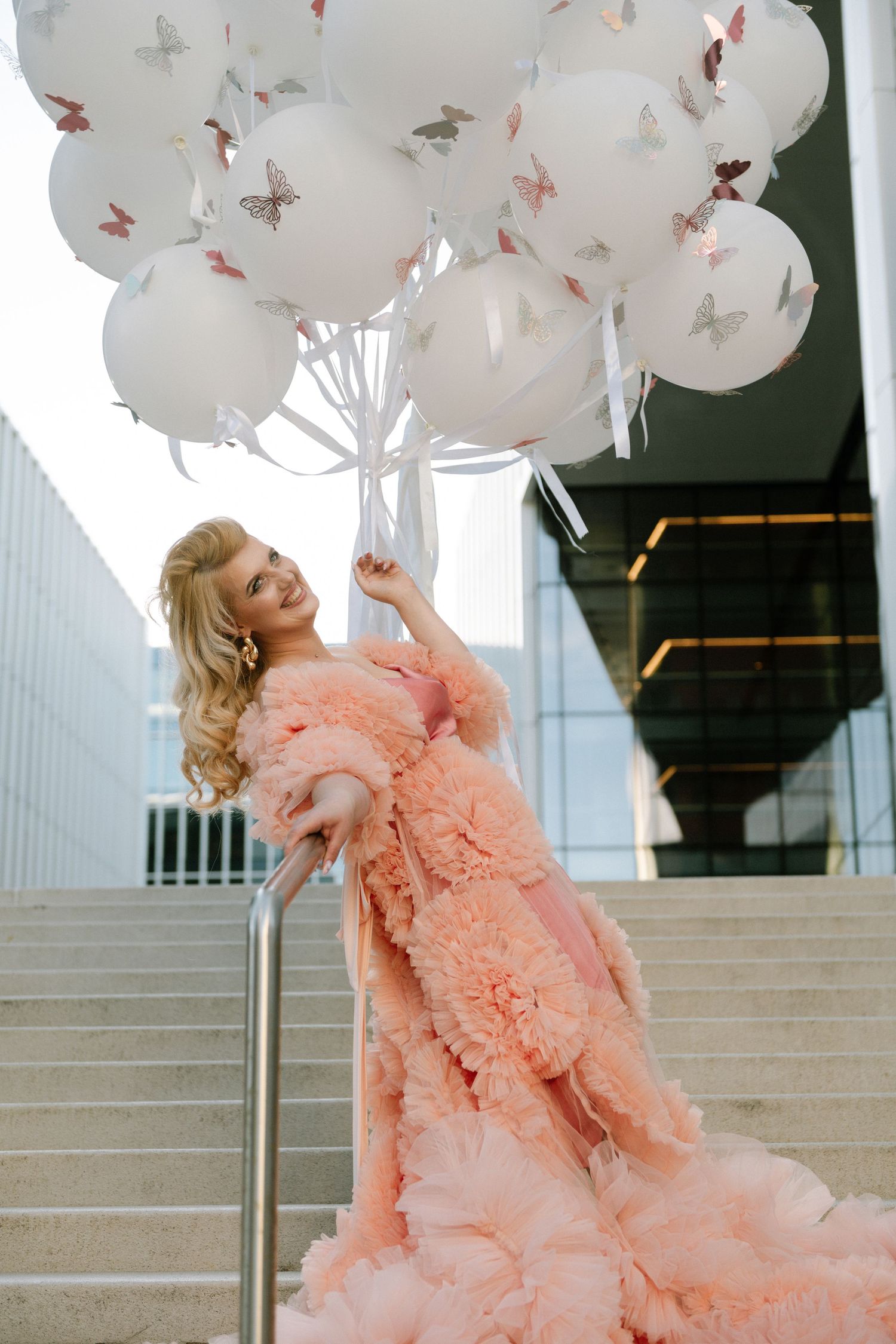 Elegant blonde woman in pink ruffled gown holding white butterfly balloons on modern building steps.