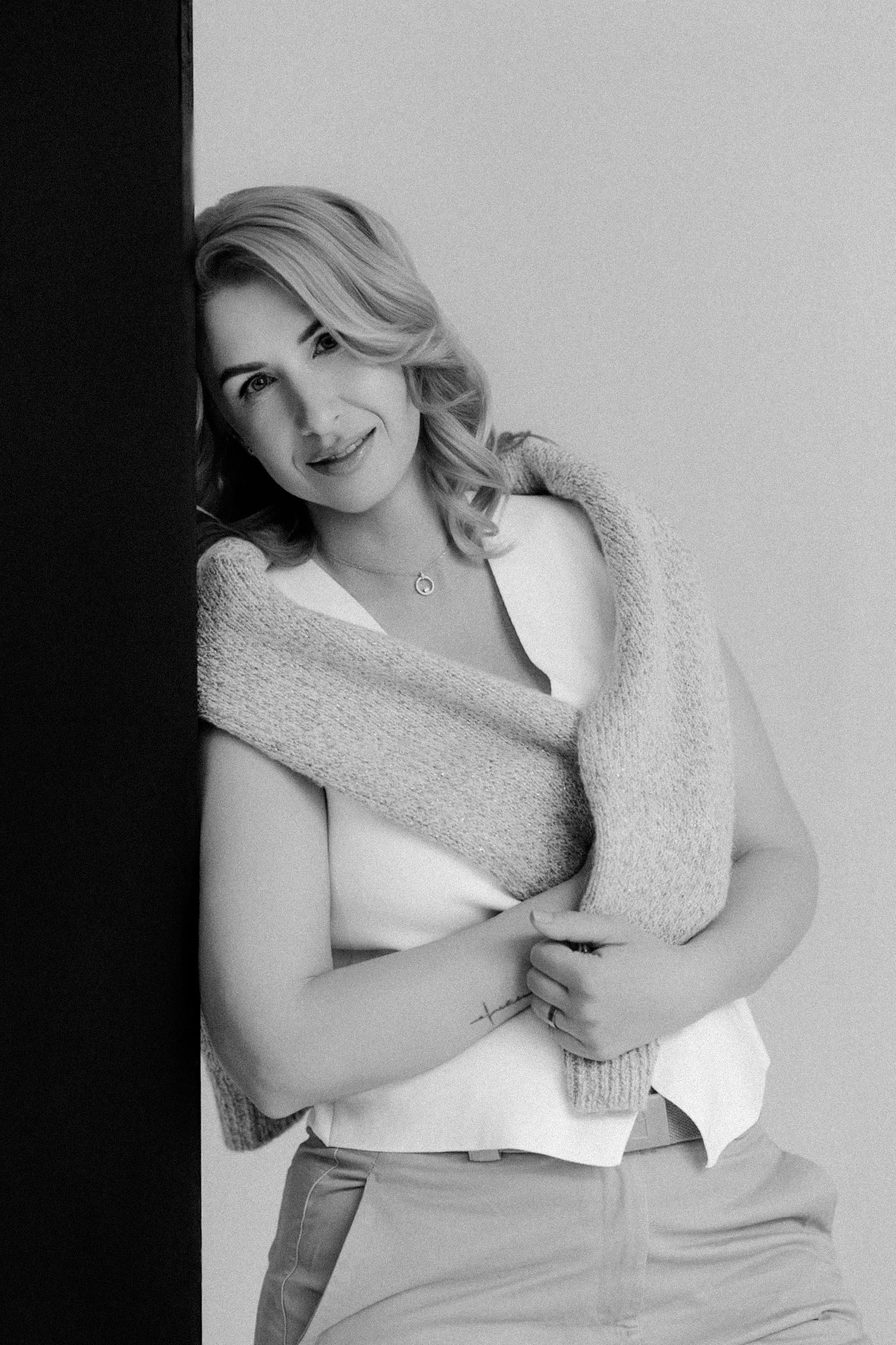 Black and white portrait of a smiling blonde woman leaning against a wall, holding a knit sweater.