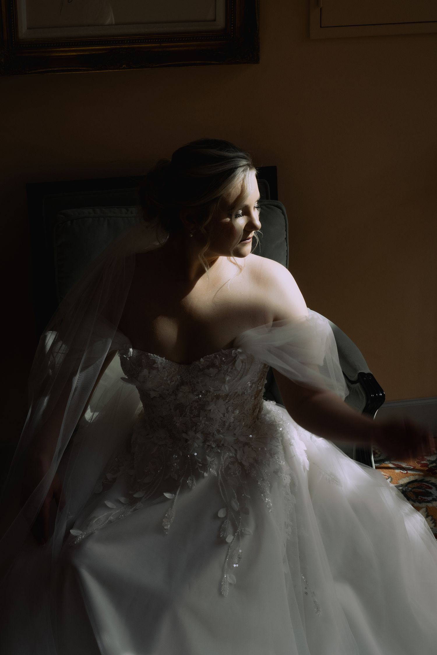 A bride in a white lace wedding dress sits in dramatic lighting creating a moody romantic atmosphere.