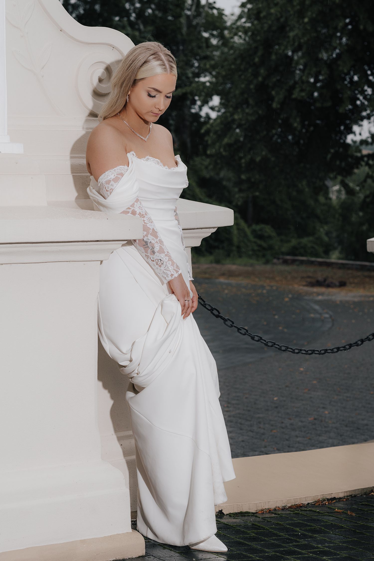 Elegant bride in off-shoulder lace sleeve wedding gown poses gracefully against white pillar in outdoor setting.