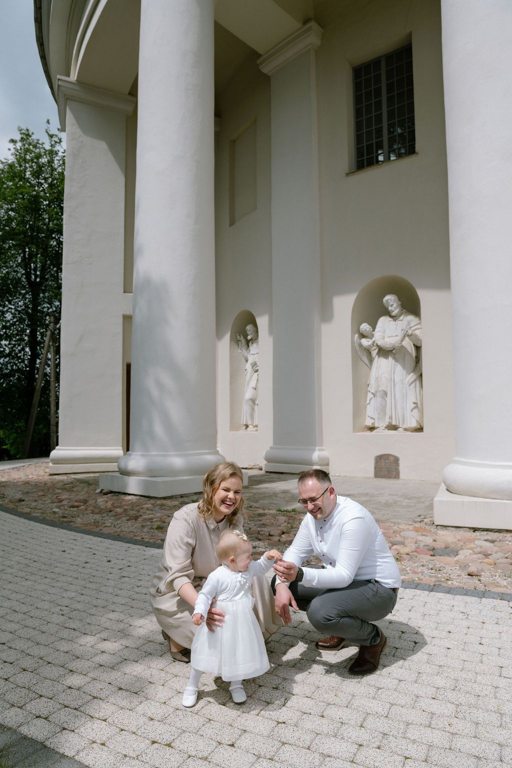 Family with baby girl in white dress posing outside a white church with religious statues and columns.