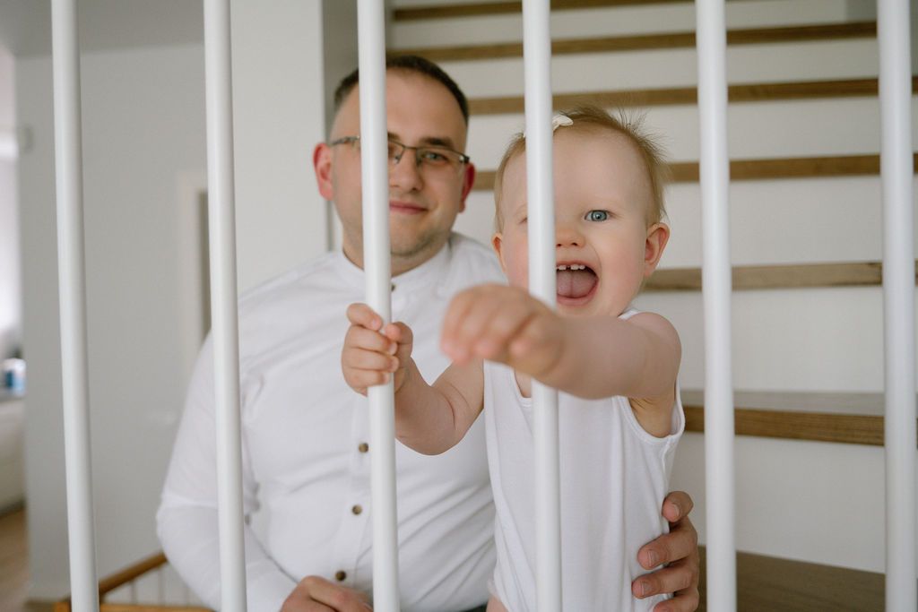 Father holding baby in white crib, smiling toddler reaching out between white wooden bars in bright nursery room.