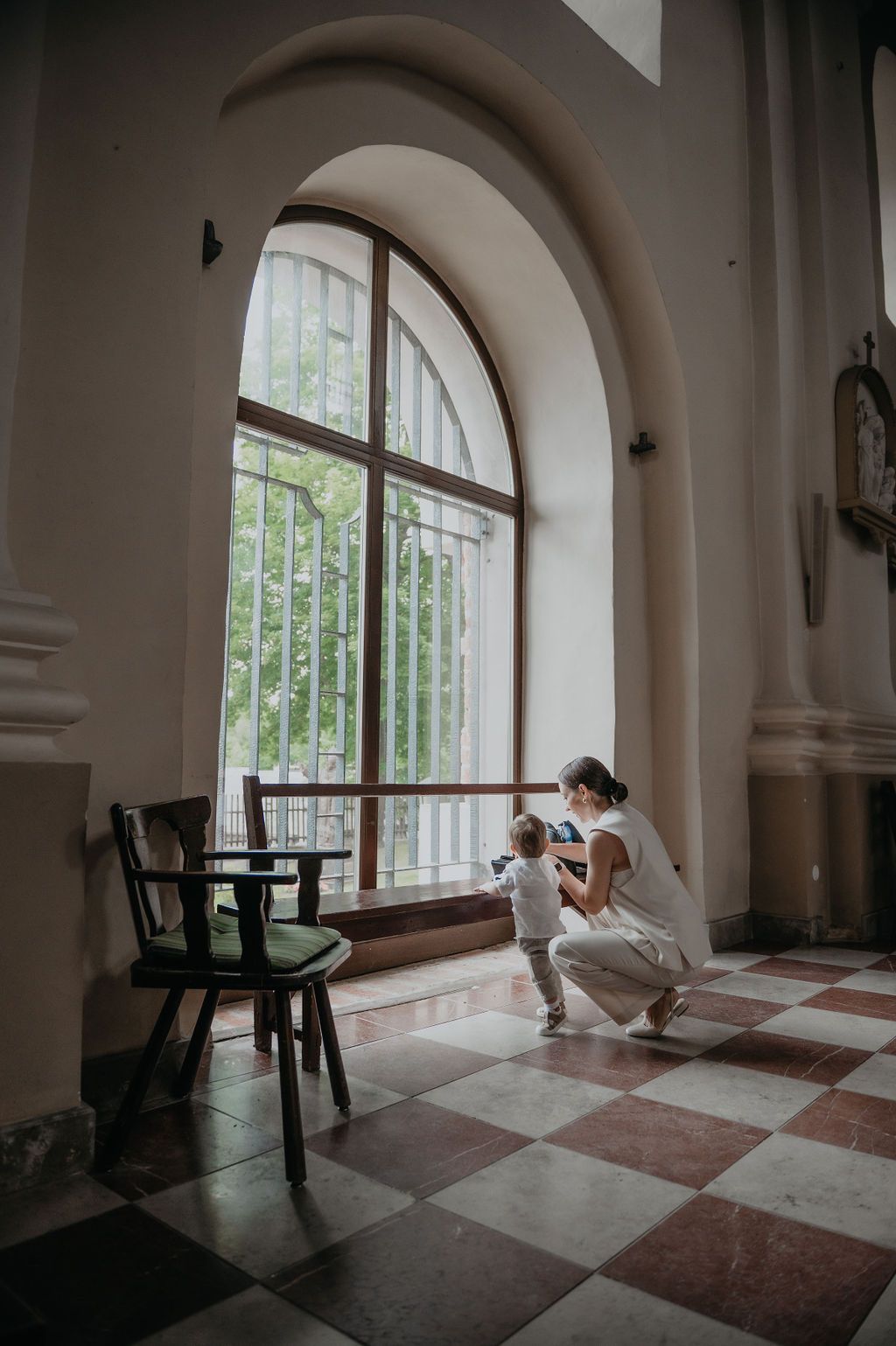Mother crouching with toddler by arched window in elegant building with checkered tile floor.