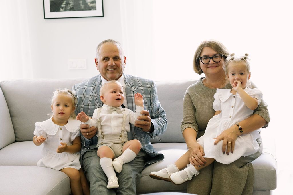 Grandparents sitting on sofa with three young children in white outfits smiling.