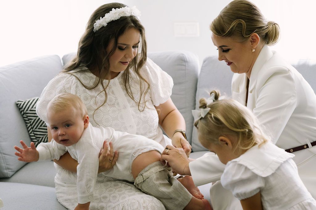 Two women in white dresses tend to a baby and toddler girl on a gray sofa during a christening ceremony.
