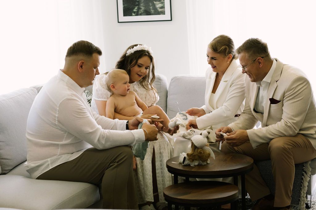 Family gathered around baby in christening outfit on sofa, sharing a tender moment with gifts and decorations.