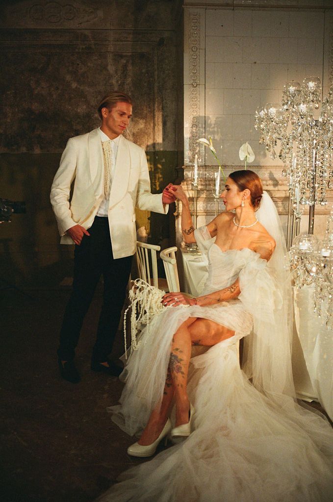 Elegant bride in white ruffled gown with veil sits as groom in white tuxedo holds her hand near crystal chandeliers.