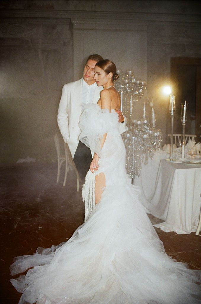 Elegant bride and groom pose in a candlelit ballroom, bride wearing a stunning backless white gown with train.