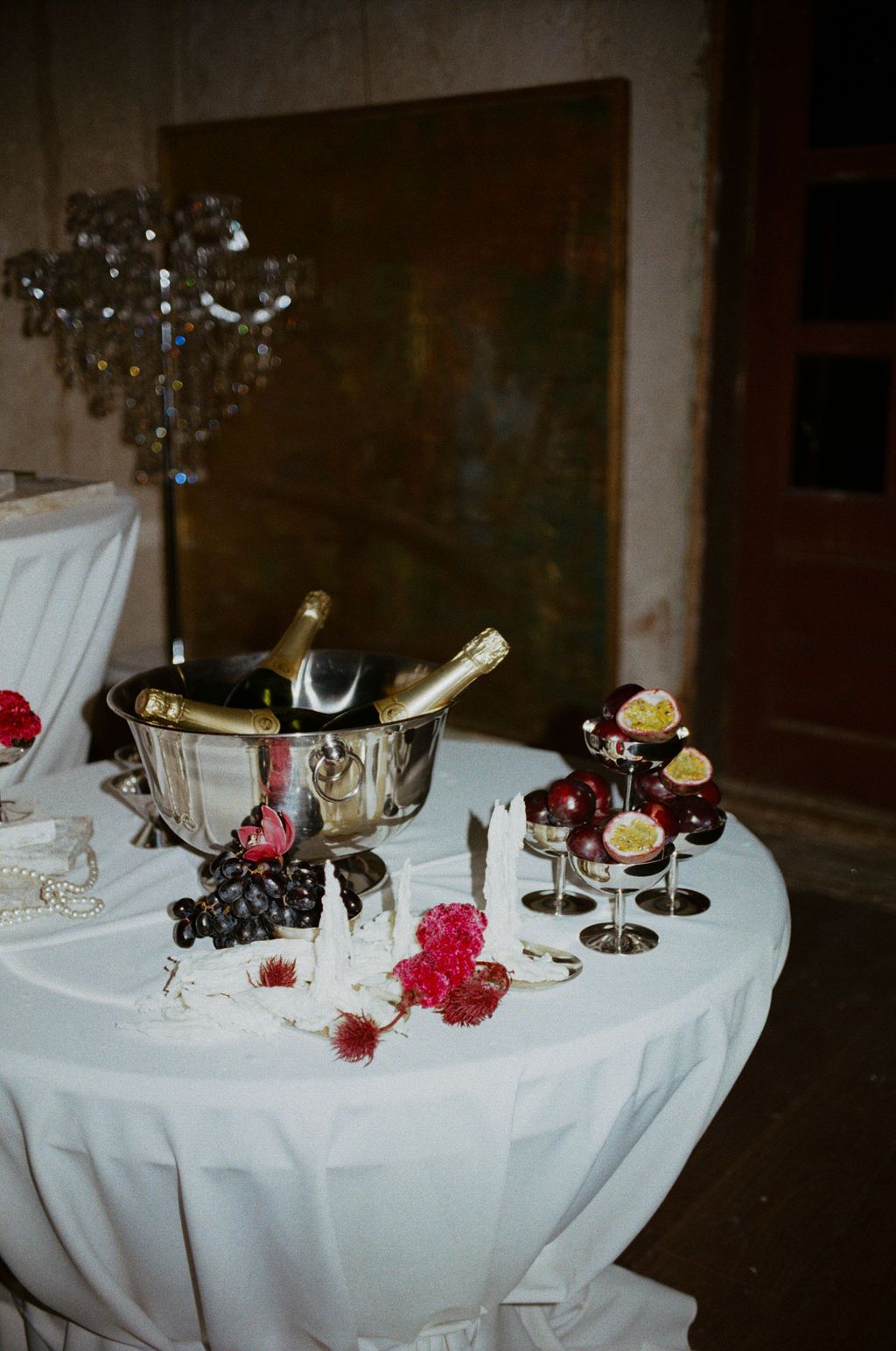 Elegant party table with champagne in ice bucket, wine glasses with citrus, grapes, and red floral decorations on white cloth.