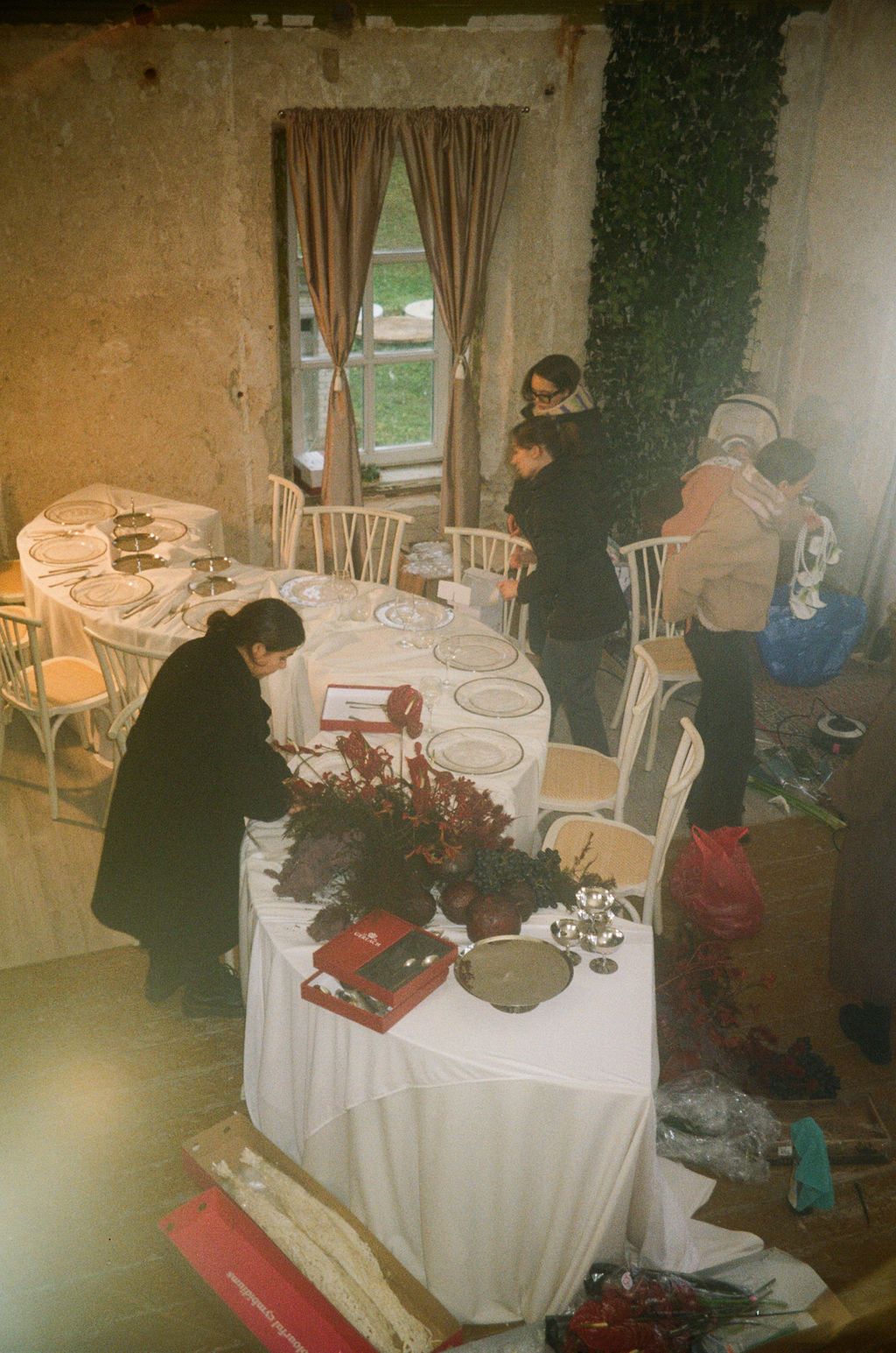 People setting up a decorated round table with white tablecloth and red floral centerpiece in a rustic stone-walled venue.