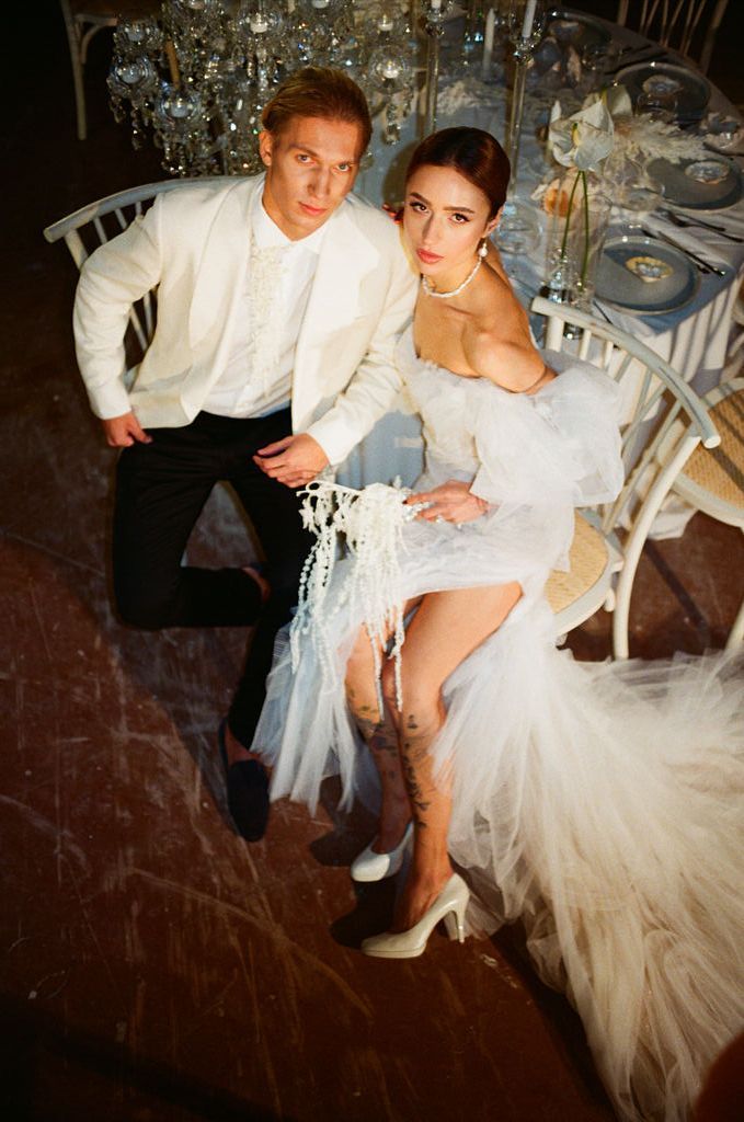 Elegant couple in white wedding attire seated together at a decorated reception table viewed from above.