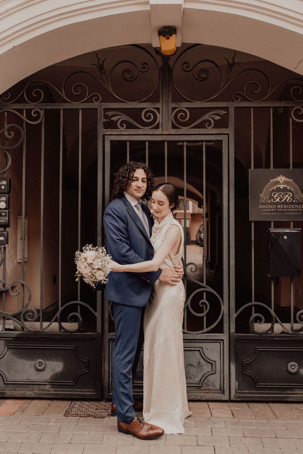 Bride and groom embrace before ornate iron gates, she holds a white bouquet in a satin gown, he wears a navy suit.