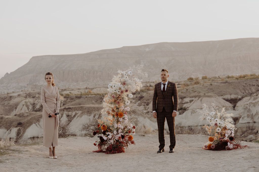 Couple stands at outdoor desert wedding ceremony with floral arrangements and rocky landscape backdrop.