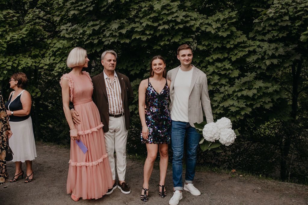 Four people posing outdoors near green trees, dressed in elegant attire, one holding white flowers.