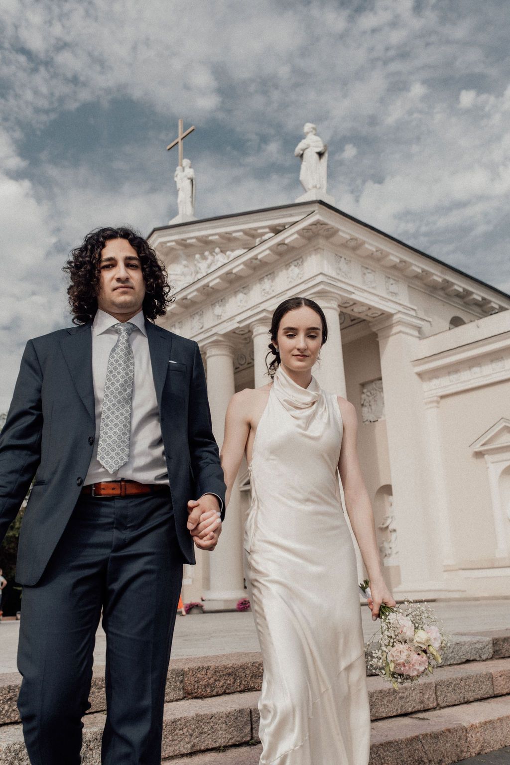 Bride and groom holding hands outside a white classical church with statues, bride in satin gown, groom in navy suit.