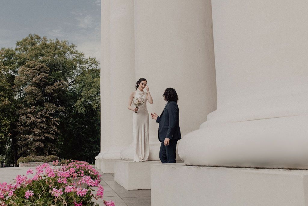 Couple holding hands in wedding attire beside white columns with pink flowers at an elegant outdoor ceremony.