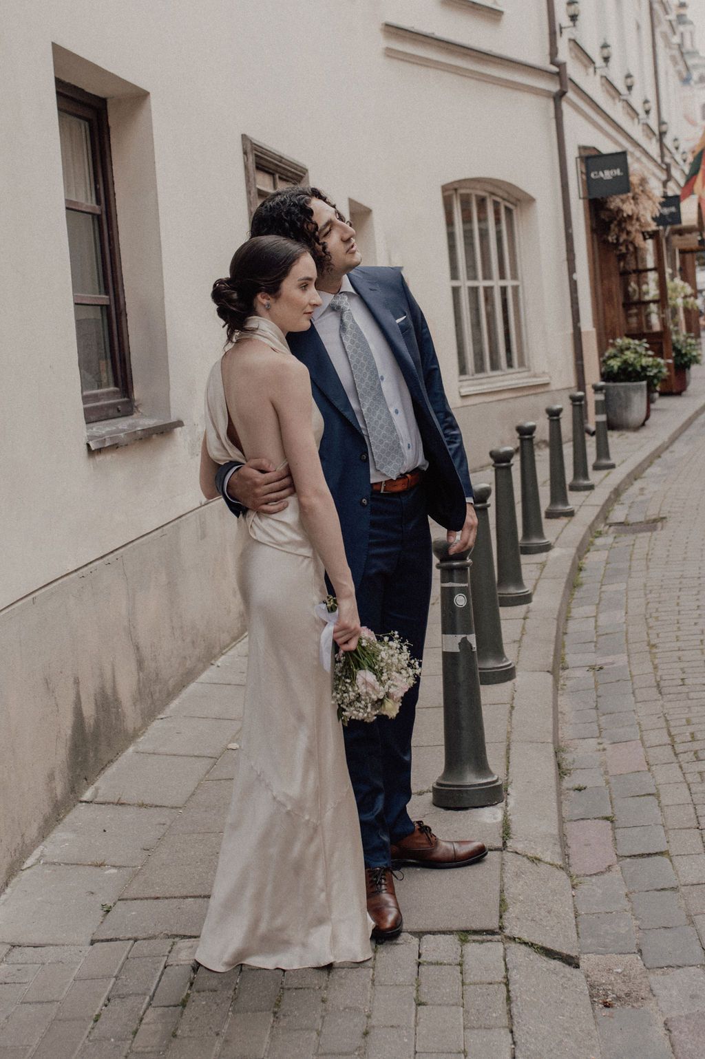 Bride in elegant backless gown holds bouquet beside groom in navy suit on cobblestone European street.