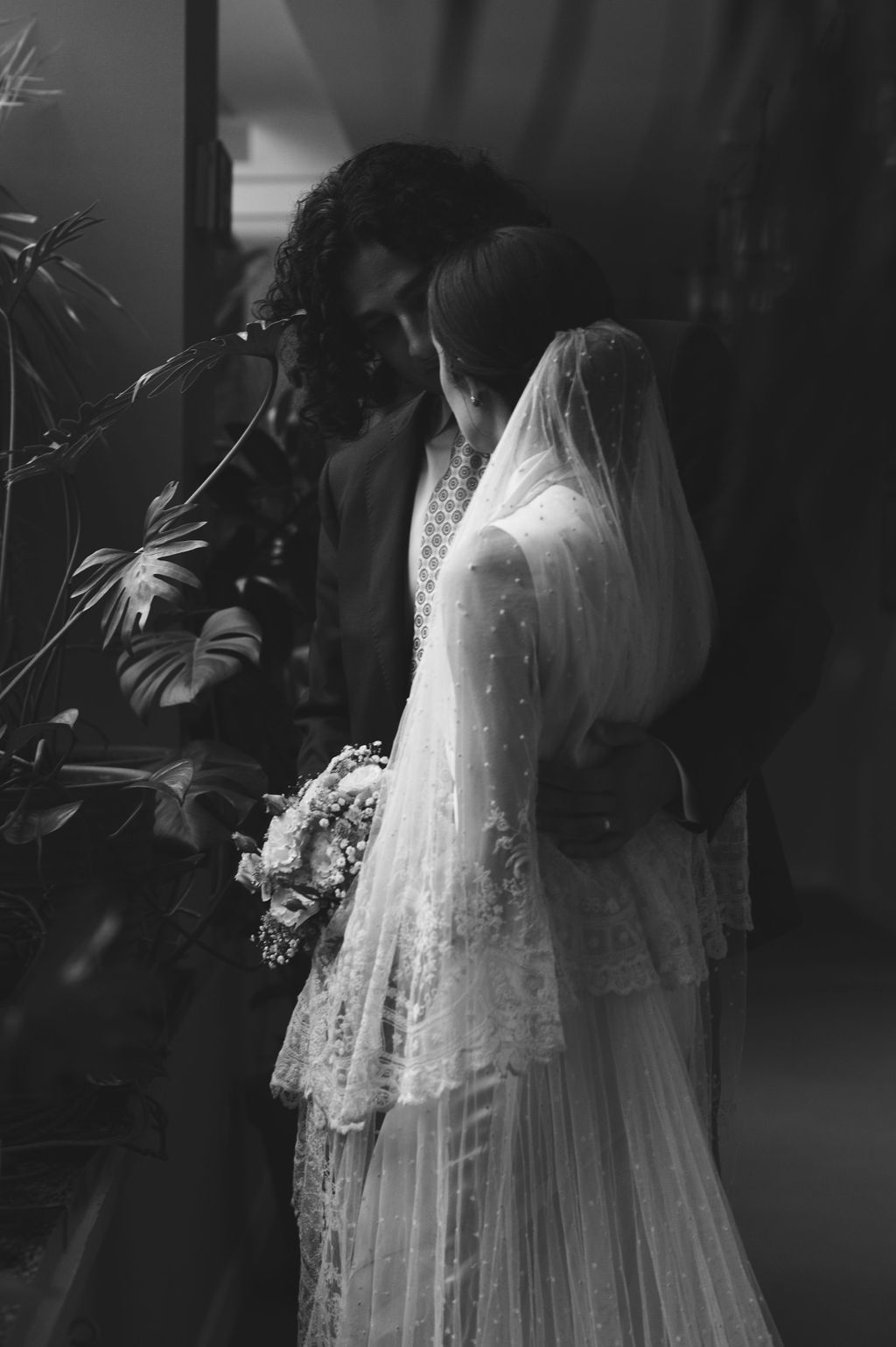 Black and white wedding photo of bride in lace gown and veil holding bouquet, sharing intimate moment with groom.