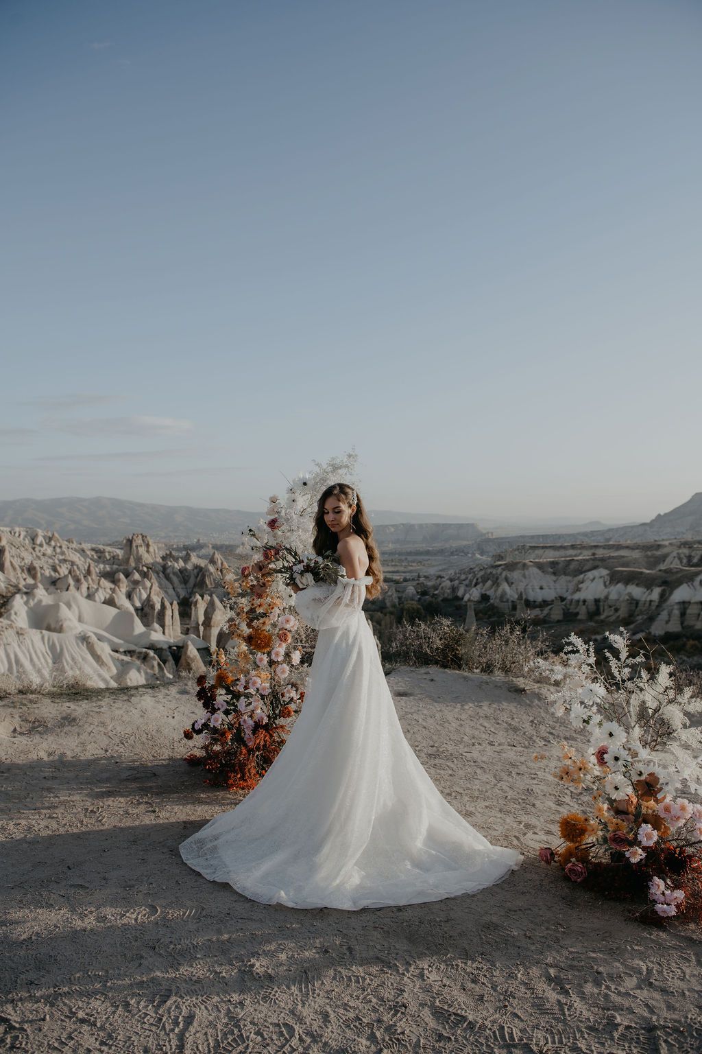 Bride in white gown poses by floral arch in Cappadocia's rocky landscape at dusk.