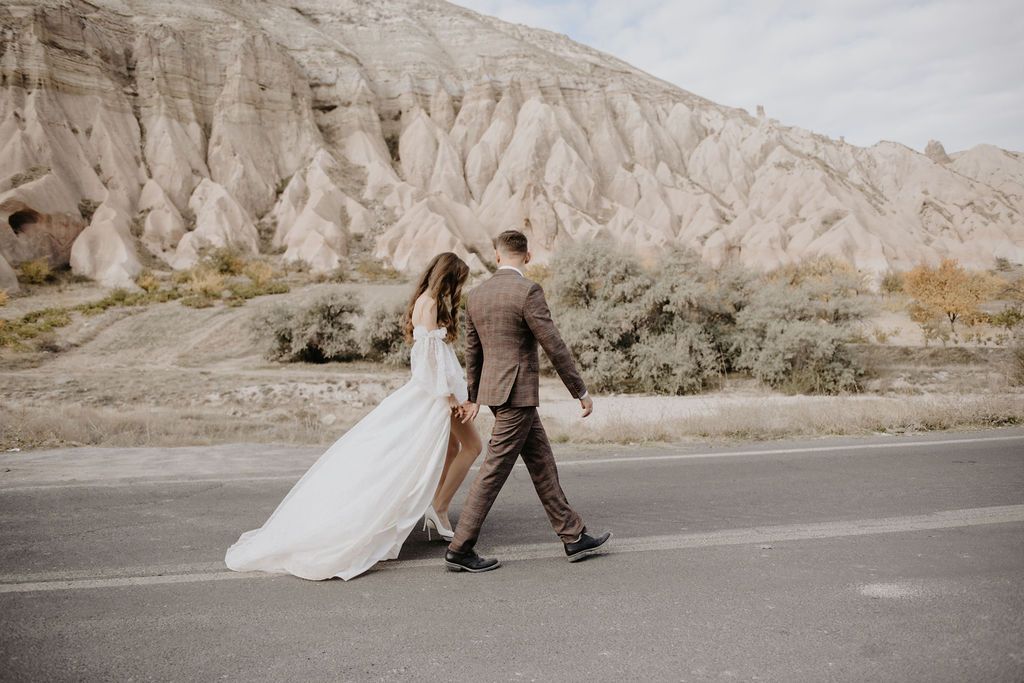 Bride and groom walking hand in hand on a road with stunning Cappadocia rock formations in the background.