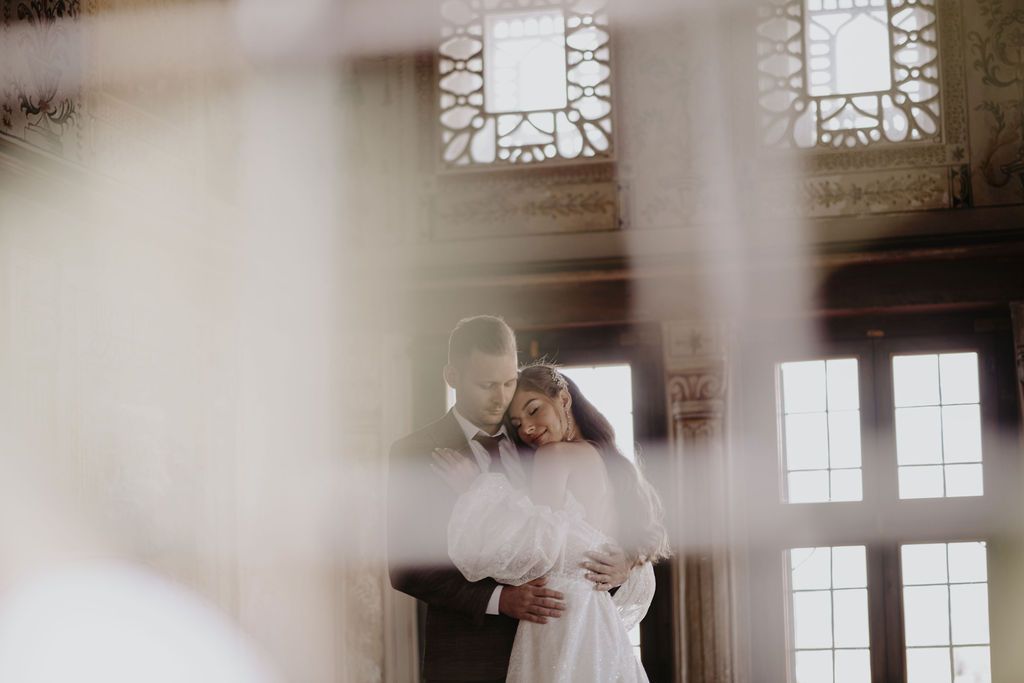 Bride and groom share romantic embrace in elegant venue with ornate windows and soft natural light on their wedding day.