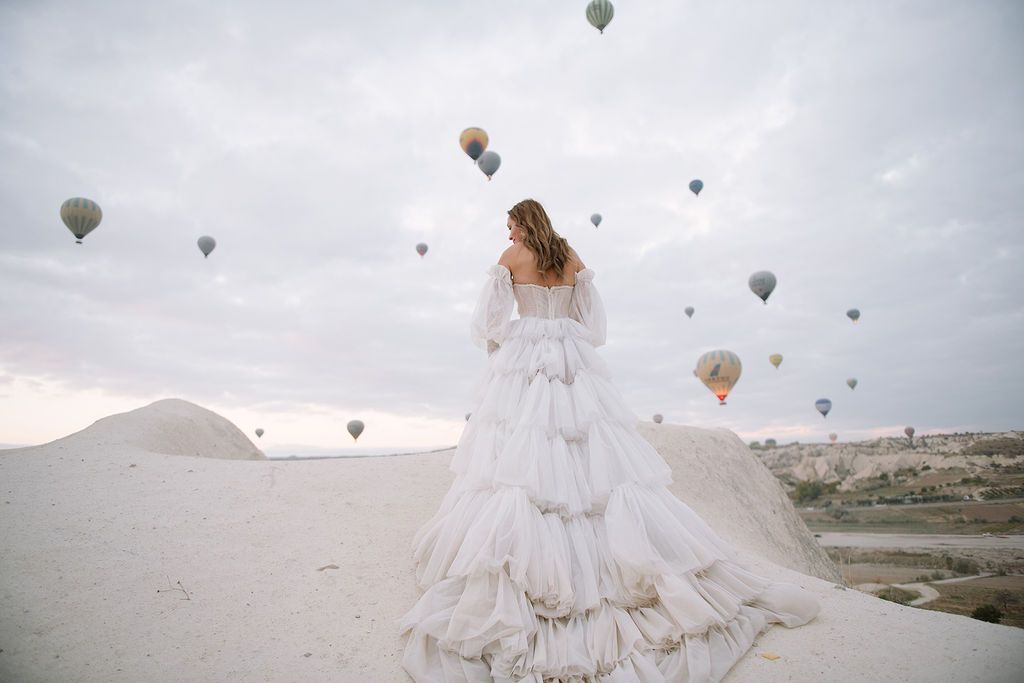 Bride in layered white gown stands on sandy dunes surrounded by colorful hot air balloons in Cappadocia.