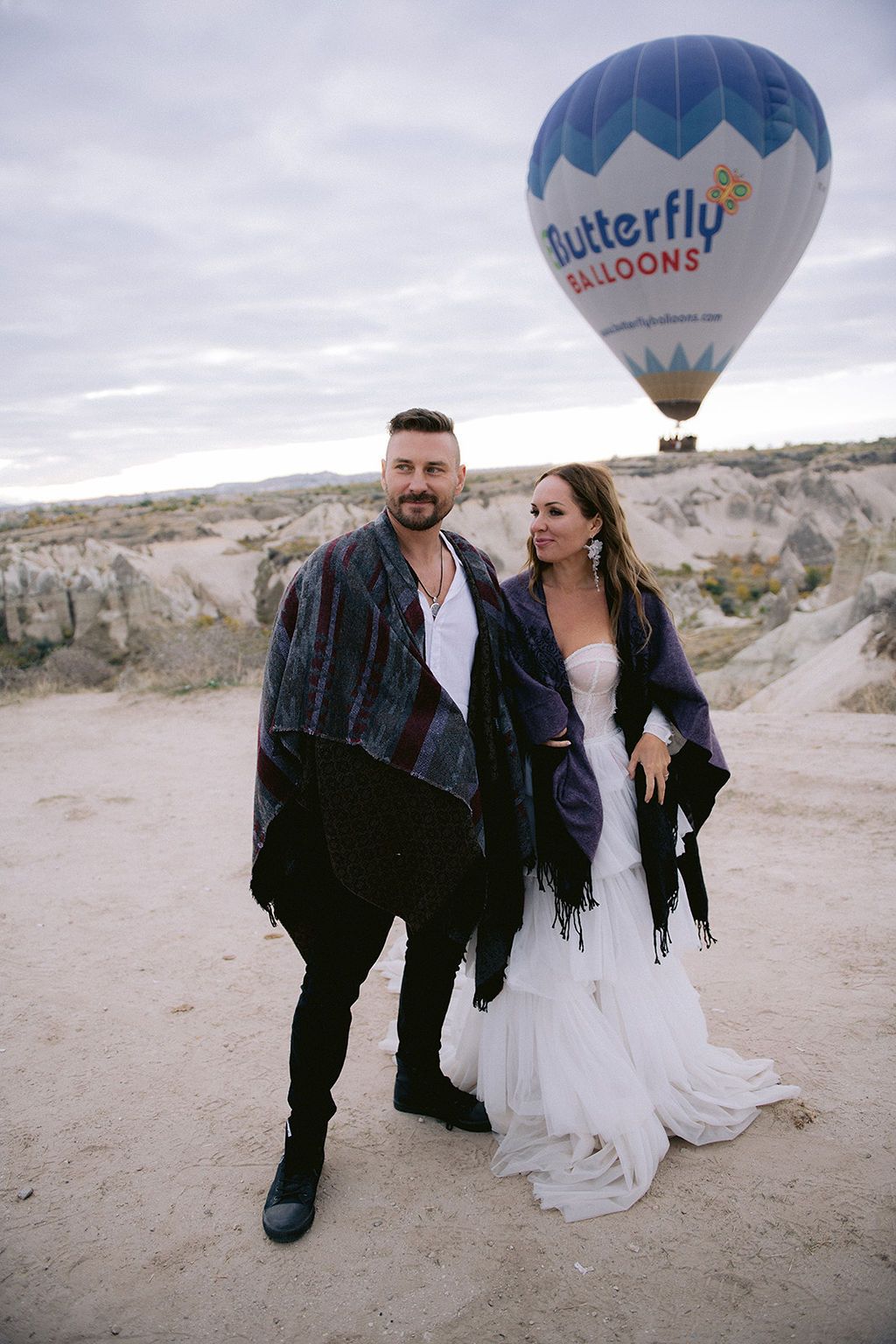 Couple in shawls poses before Butterfly Balloons hot air balloon in Cappadocia rocky landscape.