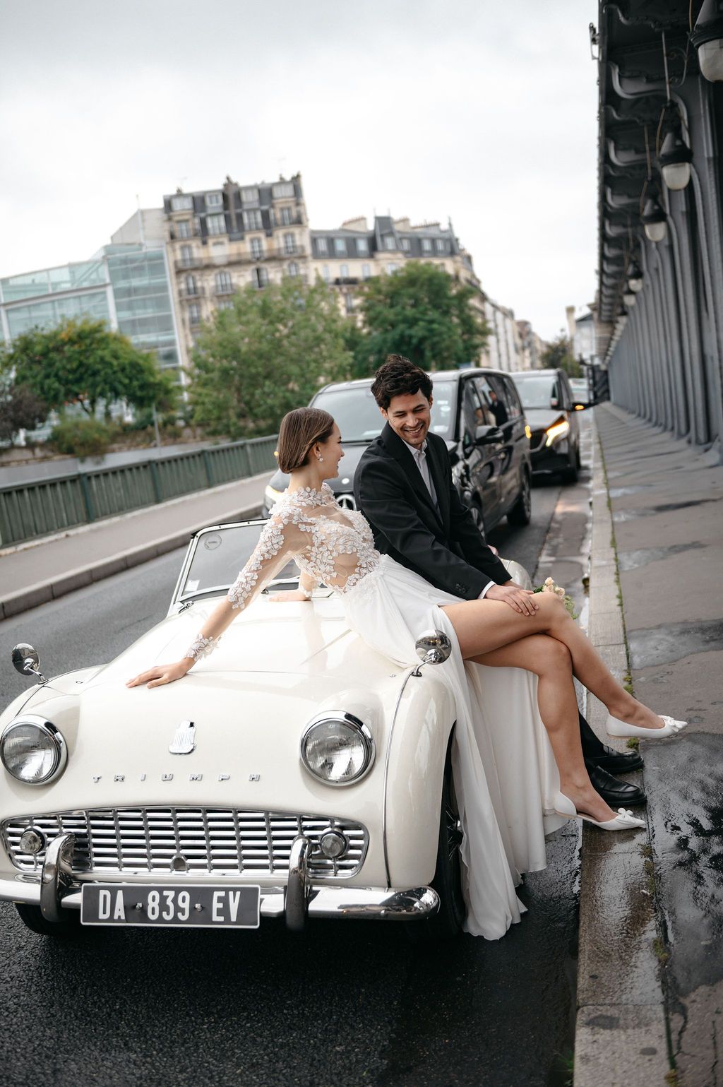 Elegant bride and groom sitting on classic white Triumph convertible on a Parisian street.