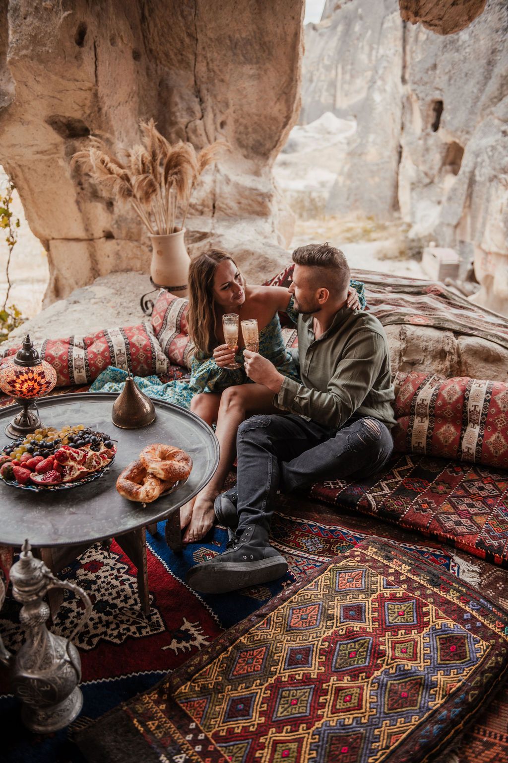 Couple toasting champagne on colorful Turkish kilim cushions with mezze spread in a cave dwelling in Cappadocia.