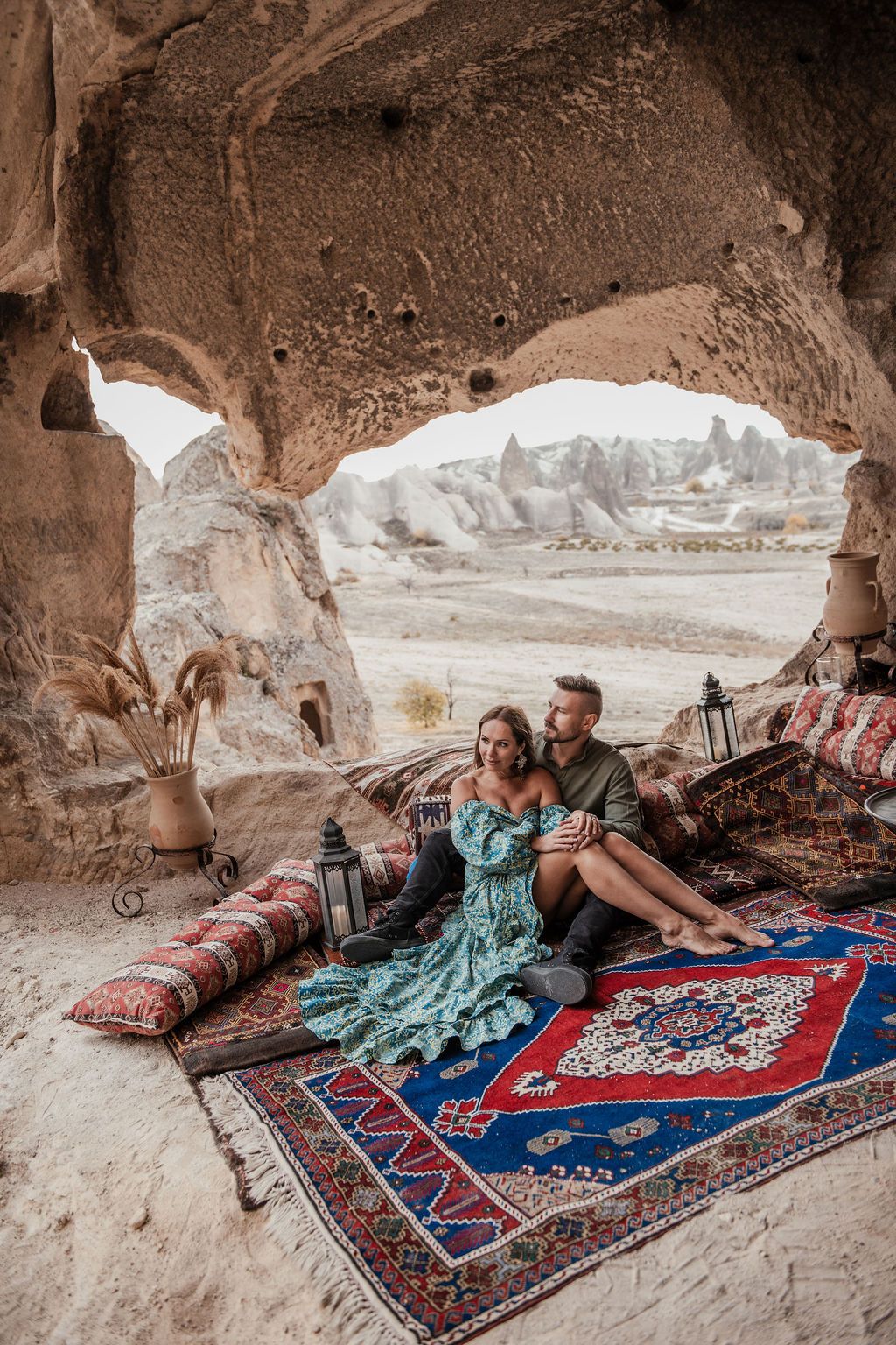 Couple relaxing on colorful rugs inside a cave dwelling in Cappadocia, Turkey, with rocky landscape views.