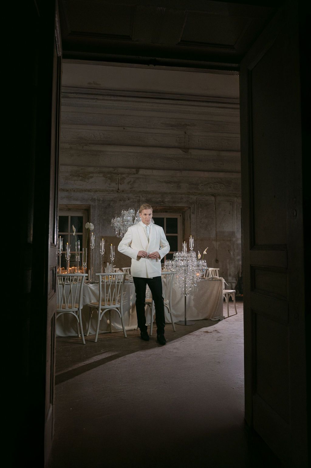 A young man in a white jacket stands in a dimly lit elegant room with chandeliers and decorated tables.