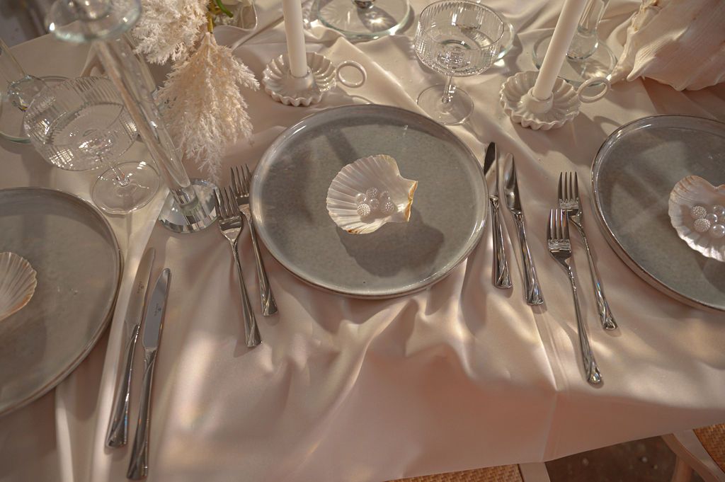 Elegant formal table setting with silver plates, crystal glasses, shell napkin holders, and polished silverware on a satin tablecloth.