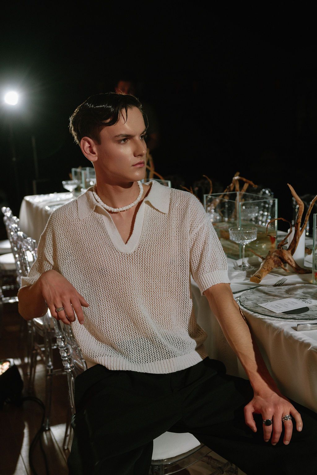 Young man in white mesh knit shirt and pearl necklace seated at elegant dinner table in dark setting.